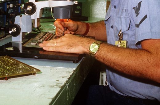 Hands performing close inspection work on a circuit board under a bench magnifier